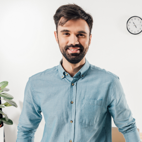Smiling businessman in a blue shirt, standing in an office setting with a plant and clock, representing professionalism in the context of BettyJet's fuel delivery services.