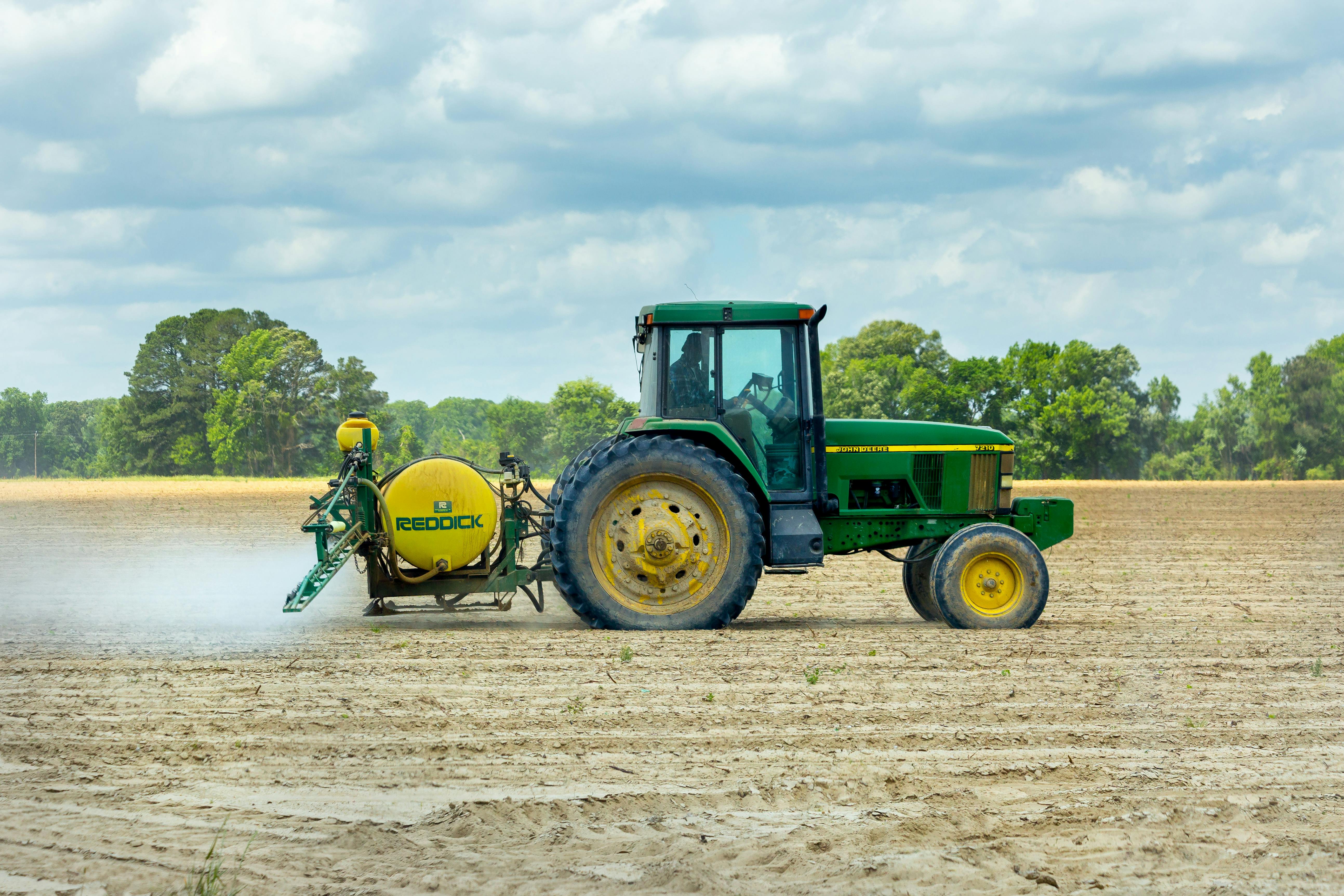 BettyJet Tractors Green tractor with yellow Reddick sprayer applying treatment to dry farmland under a cloudy sky, representing BettyJet’s diesel delivery support for agricultural operations in Florida.