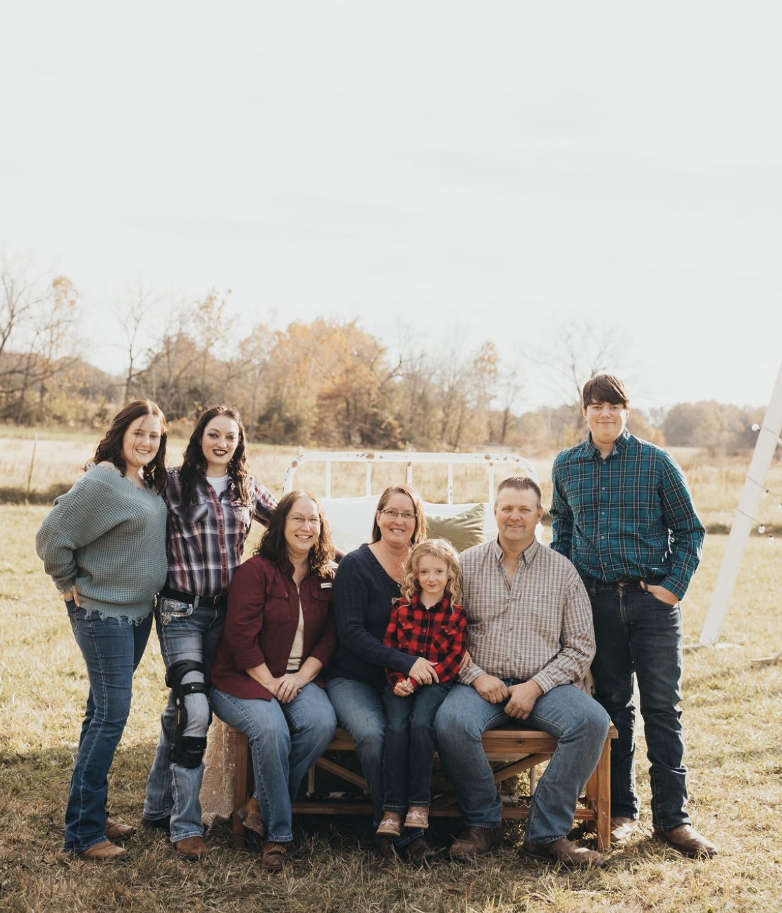 Family portrait in a rural setting, featuring seven individuals seated on a wooden bench, dressed in casual attire, with autumn foliage in the background, conveying a sense of community and warmth.